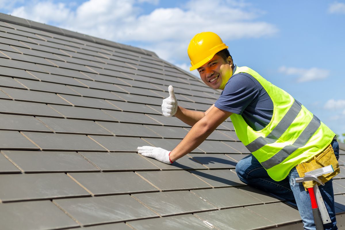 A man wearing a hard hat and safety vest stands on an asphalt shingle roof, overseeing the worksite.
